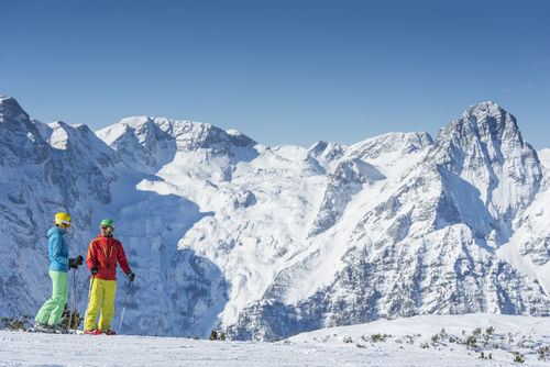 Zwei Schifahrer stehen auf der Piste, im Hintergrund die Berge