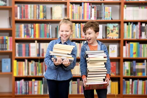 Zwei fröhliche Kinder mit Bücherstapel in Händen vor einem Regal voll mit Büchern