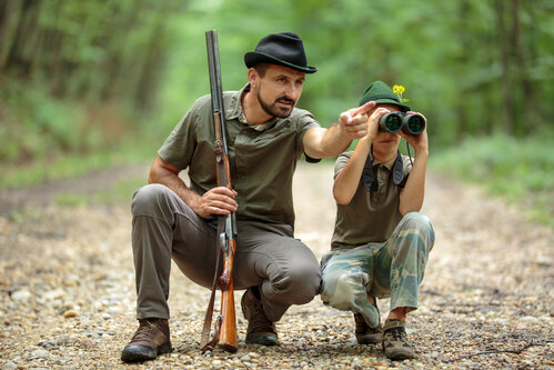 Ein Jäger, der ein Gewehr in der Hand hält und ein Junge, der durch ein Fernglas schaut, beobachten Tiere im Wald