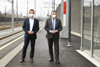 Thomas Fruhmann und Landesrat Günther Steinkellner, beide mit FFP2-Masken, auf einem Bahnsteig, im Hintergrund Gleise, Oberleitungen, Lärmschutzwand 