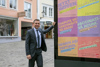 Landesrat Dr. Wolfgang Hattmannsdorfer steht in der Linzer Altstadt vor einem Plakat, auf dem in sechs verschiedenen Sprachen für die Impfung geworben wird