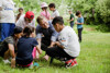 Landesrat Stefan Kaineder mit vier Kindern um ihn auf einer Wiese, alle betrachten ein Stück Erde mit Gras, im Hintergrund weitere Kinder auf der Wiese