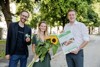 Norbert Rainer (Leiter Klimabündnis OÖ), die Gewinnerin Tamara Reiter und Landesrat Stefan Kaineder stehen neben einander im Landhauspark; Norbert Rainer hält einen kleinen Traktor, Tamara Reiter mehrere Sonnenblumen und Landesrat Stefan Kaineder einen Urlaubsgutschein in Händen 