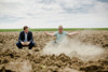 LR Kaineder und ein Mann knien auf einem trockenen Acker, vor dem Man ist eine kleine Nebelwolke aus Erde. Oberhalb befindet sich der Himmel und hinter den Personen ein Maiskornfeld.