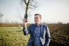LR Stefan Kaineder steht im Halbportrait auf einer Wiese, dahinter Äcker und Bäume. Er hält ein Smartphone in der Hand.