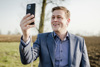 Landesrat Stefan Kaineder schaut in ein Mobiltelefon, im Hintergrund Wiese und Acker
