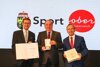 Landesrat Markus Achleitner mit Urkunde, DI Erich Haider mit Medaille und Franz Schiefermair mit Tortenschachtel in Händen stehen nebeneinander vor einer großen Videowand mit Landeswappen und Beschriftung Sport Oberösterreich