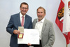 Landesrat Markus Achleitner und Johann Kaindlstorfer stehen nebeneinander und halten Urkunde und Medaille in Händen, im Hintergrund eine Oberösterreich-Fahne