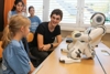 Fünf Jugendliche, zwei sitzend, drei stehen und davor auf dem Klassenzimmertisch ein sitzender Roboter.