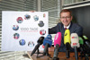 Landesrat Markus Achleitner an einem Konferenztisch mit vielen Mikrofonen, er hält ein Poster mit Beschriftung 1000 Tage Vollgas für Oberösterreich und im Kreis angeordneten Fotos mit Szenen seiner Tätigkeiten