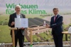 Landesrat Markus Achleitner und Herbert Ortner mit dem Goldenen Verdienstzeichen des Landes OÖ und einer Urkunde vor einem Plakat mit einer Grafik des neuen Betriebsgebäudes und der Aufschrift Wir bauen für des Klima