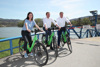 Petra Riffert, Landesrat Markus Achleitner und Friedrich Kaindlstorfer, MBA, nebeneinander sitzend jeweils auf einem Rad, auf einer Auffahrt zu einer Fähre, im Hintergrund die Donau, Ufer mit Häusern und Bäumen