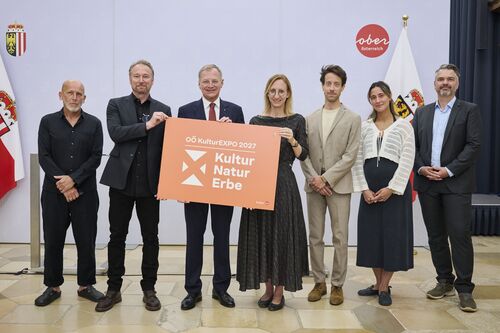 Landeshauptmann Stelzer und Landeskulturdirektorin Nazzal halten gemeinsam das Logo mit der Aufschrift „OÖ KulturEXPO 2027 Kultur – Natur – Erbe“.