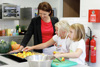 Landesrätin Michaela Langer-Weninger steht mit zwei Kindern in einer Küche bei einer Arbeitsplatte, die Kinder legen Kürbisstückchen auf ein Backblech