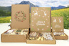 Drei verschieden große weihnachtlich bedruckte Schachteln, gefüllt mit verschiedenen Lebensmittelartikeln, liegen nebeneinander, im Hintergrund Fototapete mit Wiese und Bergen