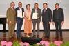 v. l. n. r.: Georg Adam Starhemberg (Obmann proHolz Oberösterreich), Roland Höfer (Simader Bau- und Zimmermeister GmbH), Sandra Gnigler und Gunar Wilhelm (mia2 ARCHITEKTUR ZT GmbH), LH-Stv. Manfred Haimbuchner, Juror Helmut Dietrich (Dietrich | Untertrifaller Architekten) stehen nebeneinander auf einer Tribüne. Davor am Boden ein Bildschirm und Blumendekoration, dahinter ein Klavier. Drei Personen halten eine Urkunde bzw. einen Preis – Skulptur in der Hand.