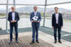 Klaus Grübl, Wirtschafts-LR Markus Achleitner - er hält eine Plakette in der Hand - und Fabian Lichtenstein stehen in einem Gebäude vor einer Glasfront, dahinter ein Fluss mit Stadt und Landschaft.