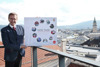 Landesrat Markus Achleitner auf einer Dachterrasse mit Blick über Linz, er hält ein Poster mit Beschriftung 1000 Tage Vollgas für Oberösterreich und im Kreis angeordneten Fotos mit Szenen seiner Tätigkeiten