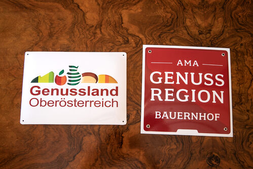 Zwei Plaketten auf einem Tisch  - „Genussland Oberösterrich“ und „AMA Genussregion Bauernhof“