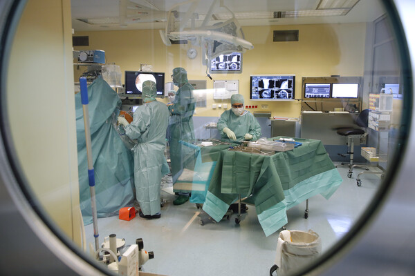 Blick durch ein rundes Fenster in einen Operationssaal, in dem mehrere medizinische Fachkräfte in grüner Schutzkleidung einen Eingriff durchführen.