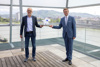 Klaus Grübl und Wirtschafts-LR Markus Achleitner stehen in einem Gebäude vor einer Glasfront, dahinter ein Fluss mit Stadt und Landschaft. Sie halten eine Plakette in den Händen.