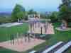 Großer Spielplatz mit Kletter- und Balanciergeräten aus Holz in einem parkartigen Gelände, weiter Blick ins Land