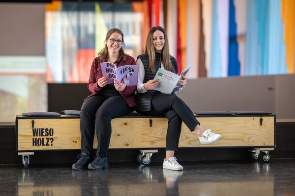 Zwei junge Frauen sitzen lächelnd auf einer langen Holzkiste und halten aufgeklappte Broschüren in den Händen. Links ist auf der Kiste der Schriftzug „WIESO HOLZ?“ zu sehen; im Hintergrund unscharfe, bunte Vertikalen in einem Innenraum.
