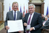 Landeshauptmann Mag. Thomas Stelzer mit Urkunde und Dr. Johann Kalliauer mit Medaille stehen nebeneinander im Steinernen Saal des Linzer Landhauses, im Hintergrund Publikum und Fahnen