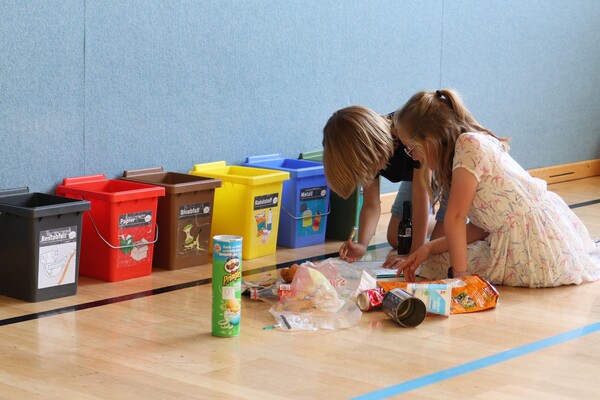 Zwei Kinder sitzen auf dem Boden und füllen etwas auf einem Blatt Papier aus, neben ihnen leere Verpackungen, hinten sind verschiedenfarbige Abfalleimer (Restabfall, Papier, Bioabfall usw.)