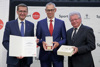 Landesrat Markus Achleitner mit Urkunde, Dr. Richard Hargassner mit Medaille und Hans Friedinger mit Tortenschachtel in Händen stehen nebeneinander vor einer Fotowand mit Beschriftung Sport Oberösterreich