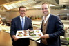 Landesrat Markus Achleitner und Manfred Kröswang stehen nebeneinander an der Theke einer Großküche und halten gemeinsam ein Tablett mit kleinen Portionen an Lebensmittel-Spezialitäten