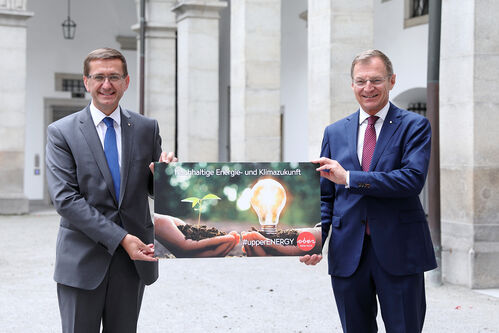 Landesrat Markus Achleitner und Landeshauptmann Thomas Stelzer im Innenhof des Landhauses, sie halten das Plakat zum Thema upperEnergy. Das Plakat zeigt zwei Hände mit Erde, einer Pflanze und einer Glühbirne.