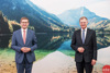 Landesrat Markus Achleitner und Landeshauptmann Mag. Thomas Stelzer stehen vor einem großen Foto, auf dem ein See abgebildet ist sowie hohe bewaldete Berge