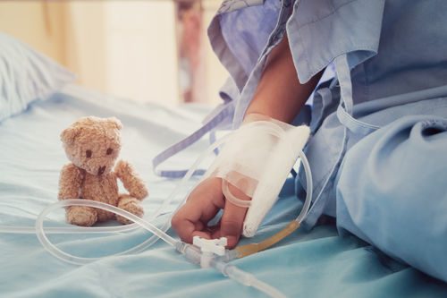 Hand eines kleinen Kindes mit Infusionsschlauch und ein Teddybär am Krankenbett.