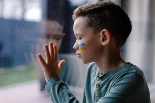 Ein Kind mit einer blau-gelben Flagge an der Wange sitzt vor einer Glasscheibe, auf die es seine Hand hält.