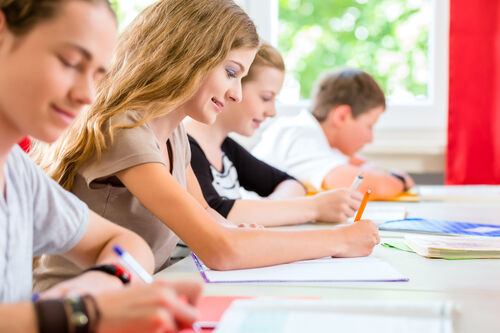 Schülerinnen und Schüler sitzen nebeneinander an einem Tisch und schreiben etwas in ihre Hefte.