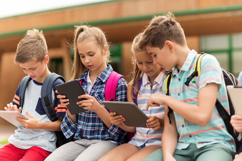 Vier Schulkinder sitzen nebeneinander, drei davon haben ein Tablet in der Hand, im Hintergrund ein Schulgebäude