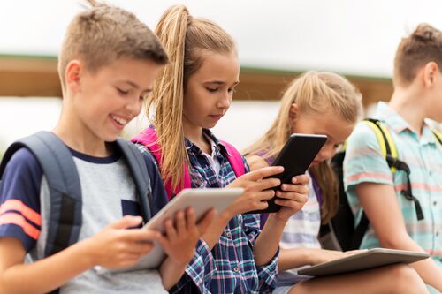 Vier junge Menschen sitzen nebeneinander vor einem Schulgebäude, jedes Kind schaut auf ein Tablet