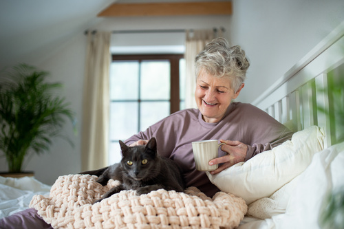 Eine ältere Frau sitzt entspannt im Bett, hält eine Tasse in der Hand und streichelt eine schwarze Katze auf der Decke.