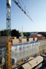 Baustelle mit Arbeitern auf einem Stadtgebäude, Kran hebt ein Holzbauteil auf den Dachausbau aus Holz, daneben weitere Stadtgebäude, im Hintergrund Baumkronen und weitere Gebäude