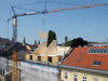 Baustelle auf einem Stadtgebäude, Kran hebt ein Holzbauteil auf den Dachausbau aus Holz, daneben weitere Stadtgebäude, im Hintergrund Baumkronen und Kirchturmspitzen