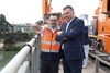 Dipl.-Ing. Stefan Dobler und LR Steinkellner stehen nebeneinander auf dem Gehsteig einer Brücke neben dem Brückengeländer , im Hintergrund ein Spezial-Lastkraftfahrzeug