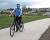 Landesrat Mag. Günther Steinkellner mit Radhelm auf einem geschotterten Radweg, im Hintergrund Wiese, Häuser