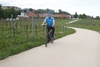 Landesrat Mag. Günther Steinkellner mit Radhelm auf einem Fahrrad auf einem Radweg, hinter bzw. neben ihm Weingärten und eine Siedlung
