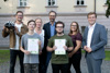 Gruppenfoto im Park vor dem Linzer Landhaus, Ulli Singer, Norbert Rainer, Beate Dunzinger und Landesrat Stefan Kaineder mit zwei Lehrlingen, die Urkunden in Händen halten, und einem Mitarbeiter, der einen Radhelm hoch hält