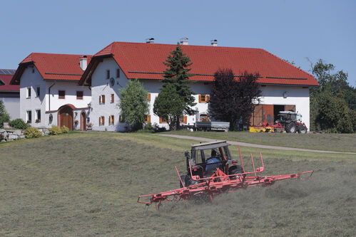 Traktor mit Heuwendemaschine auf gemähter Wiese, im Hintergrund großer Bauernhof und ein weiterer Traktor