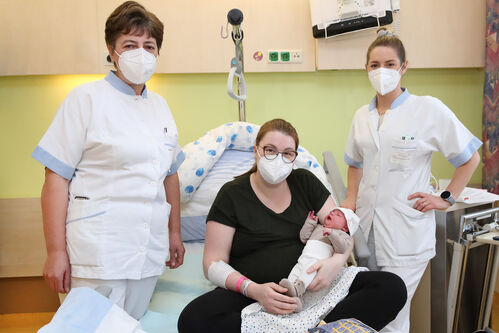 Frau in einem Krankenhausbett mit einem Neugeborenen, flankiert von zwei Krankenschwestern
