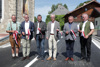 Straßenmeister Johann Strasser von der Straßenmeisterei Uttendorf, Bürgermeister Erich Rippl, LAbg. David Schiessl, Abteilungsleiter Straßenneubau und -erhaltung Christian Dick, Dechant Marek Michalowski und Diakon Martin Muigg stehen nebeneinander auf einer Straße neben einer Kirche; jeder von ihnen hält ein Stück des durchgeschnittenen Bandes in ihren Händen.