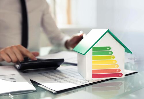 Mann sitzt an einem Tisch und tippt in einen Rechner, auf dem Tisch Unterlagen mit Zahlenreihen und das Modell eines Hauses, auf dem Energiewerte in Form verschiedenfarbiger Pfeilen symbolisiert sind