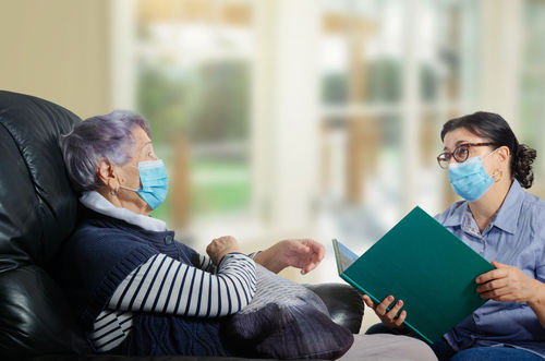 Ältere Frau mit sitzt einer jüngeren Frau, die ein Buch hält, gegenüber, beide tragen einen Mund-Nasen-Schutz